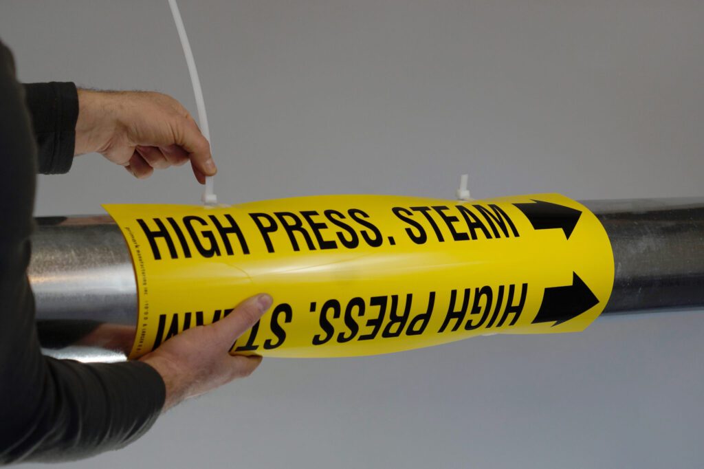 A man demonstrates how to apply the tie strap marker to a metal pipe. The man is pulling a zip tie to tighten the grip around the pipe. The marker is yellow with black text legend reads high pressure steam with an that points to the right