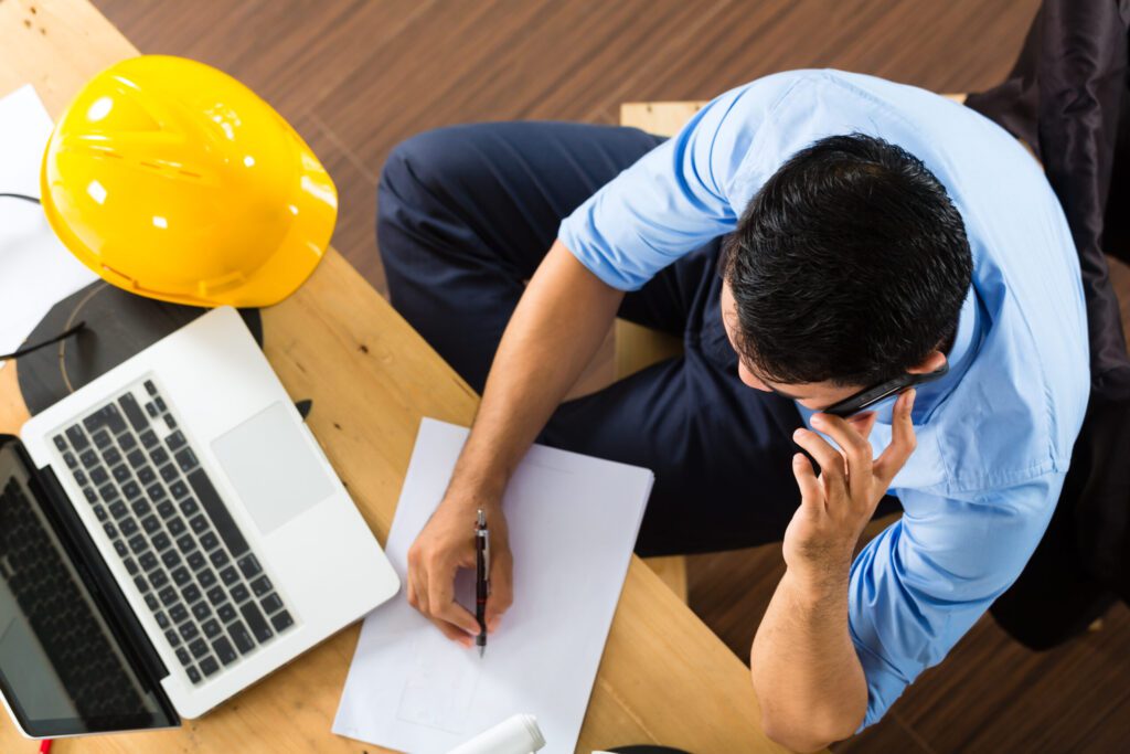 A top down View of a man in front of a desk riding notes beside a laptop and a hard hat while he's on the phone