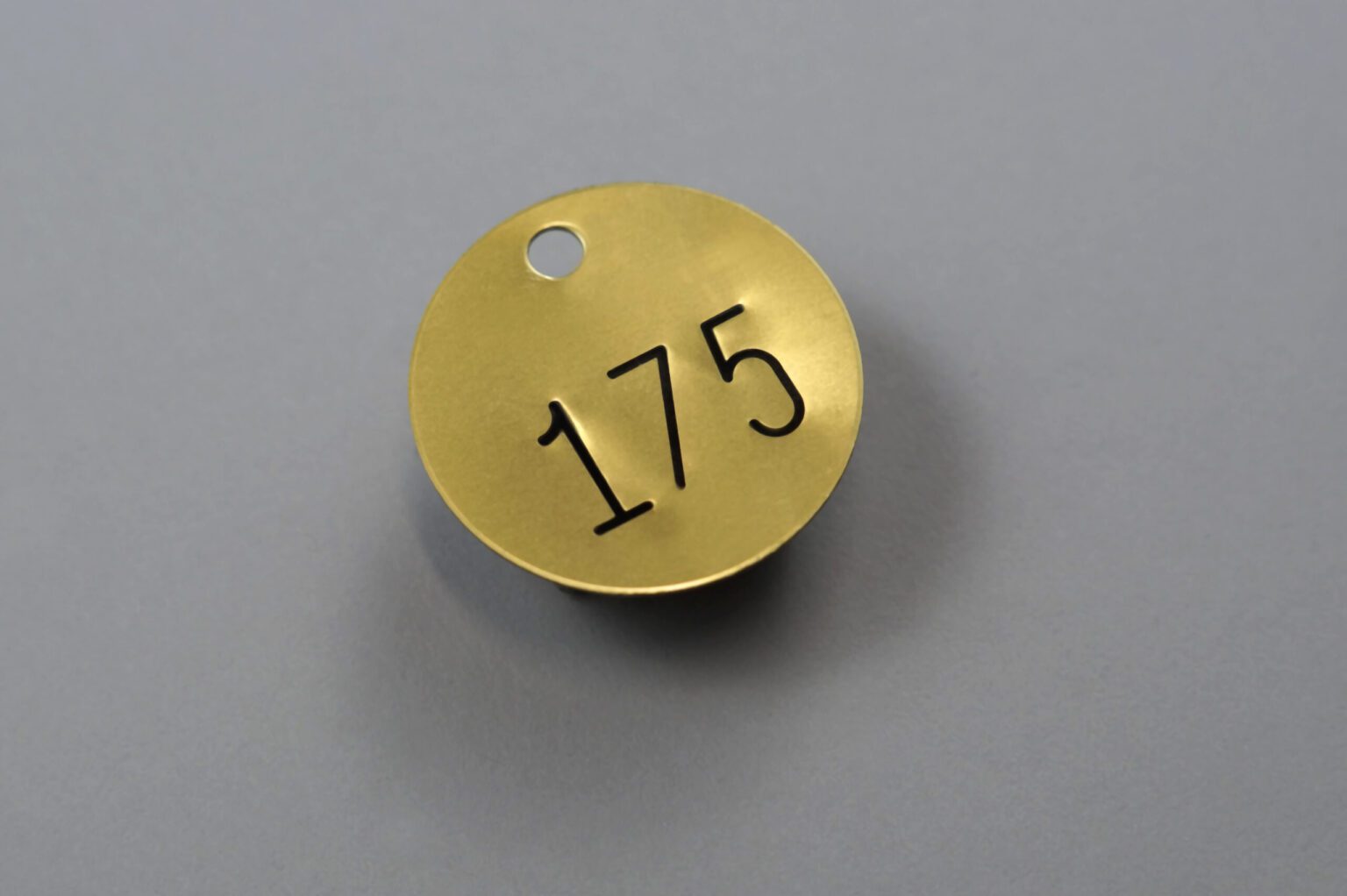 A golden circular valve tag with black text that reads "175" with a small hole at the top. On a clean gray background